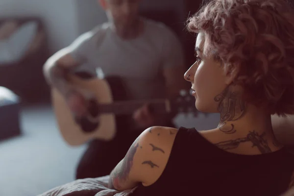 Парень играет на акустической гитаре для своей татуированной девушки дома — стоковое фото