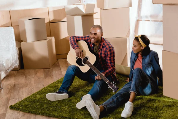 Homme afro-américain jouer sur la guitare acoustique pour petite amie dans un nouvel appartement avec des boîtes en carton — Photo de stock