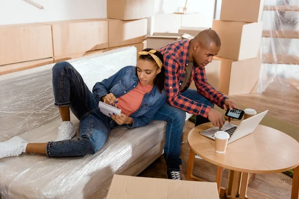 Jeune couple afro-américain planification avec bloc-notes et gadgets sur le canapé dans une nouvelle maison — Photo de stock