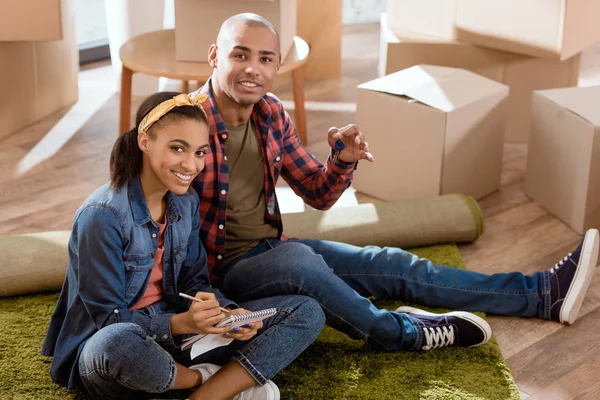 Pareja afroamericana con llaves y bloc de notas sentado en nuevo apartamento - foto de stock