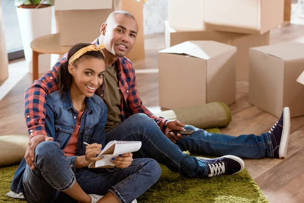 Sonriente pareja afroamericana con smartphone y bloc de notas sentado en nuevo apartamento - foto de stock