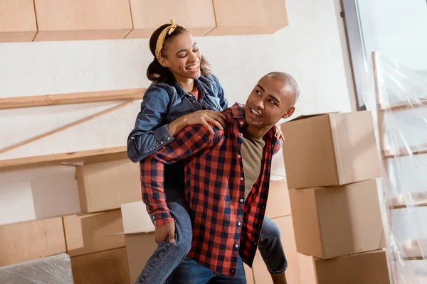 Feliz africano americano pareja piggybacking en nuevo apartamento con cajas de cartón - foto de stock