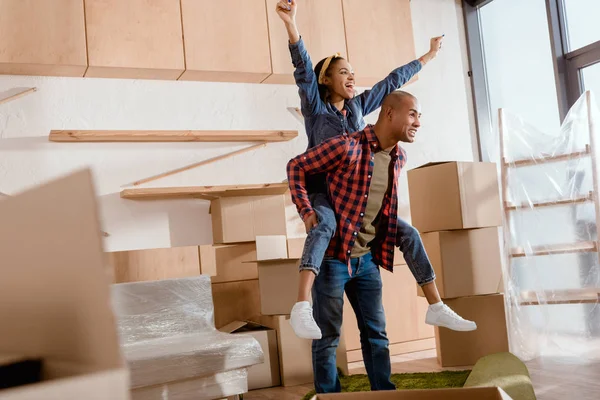 Excited african american couple piggybacking in new apartment with cardboard boxes — Stock Photo