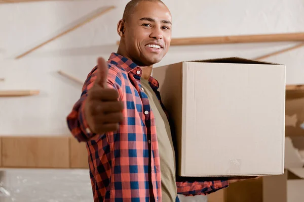 Young african american man with thumb up holding cardboard box in new home — Stock Photo