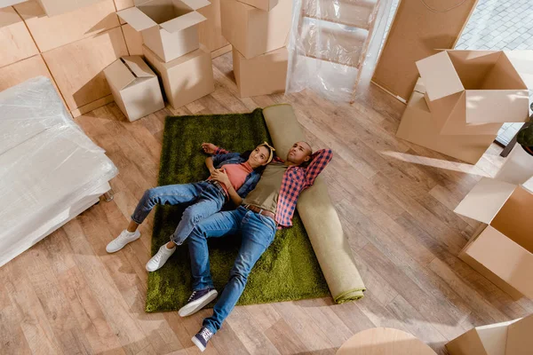 Vue de dessus du couple américain africain rêveur couché sur le sol dans une nouvelle maison avec des boîtes en carton — Photo de stock