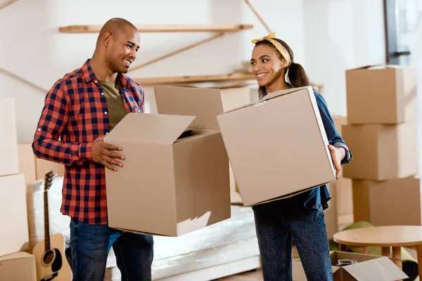 Sonriente pareja afroamericana sosteniendo cajas de cartón y mudándose a un nuevo hogar - foto de stock