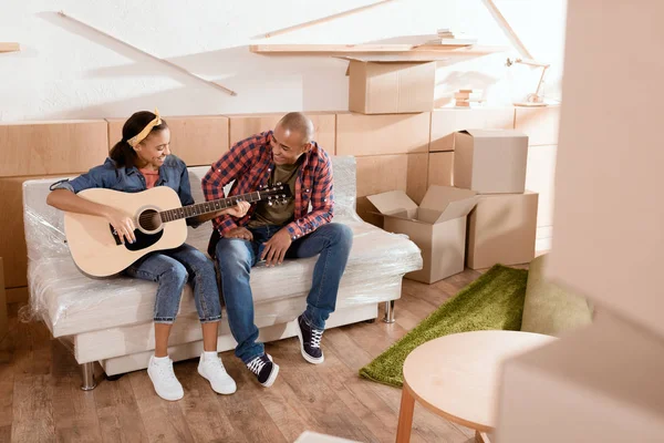 Casal afro-americano tocando guitarra acústica em nova casa com caixas de papelão — Fotografia de Stock