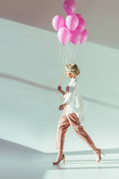 Vista lateral de hermosa mujer joven con estilo caminando con globos de color rosa en gris - foto de stock