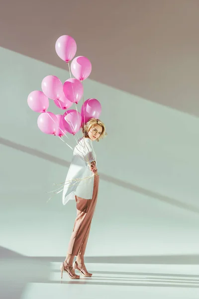 Hermosa mujer joven de moda sosteniendo globos de color rosa y mirando a la cámara en gris - foto de stock