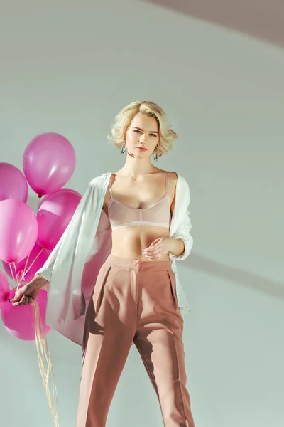 Hermosa mujer joven con ropa elegante y sujetador sosteniendo globos de color rosa y mirando a la cámara en gris - foto de stock