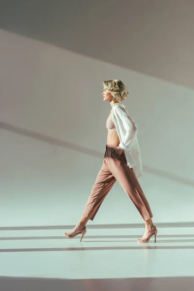 Seitenansicht der stilvollen blonden Frau in rosa BH, Hemd und Hose, die im Studio auf grau läuft — Stockfoto