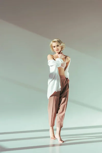 Full length view of beautiful barefoot blonde woman in pink bra, shirt and pants looking at camera on grey — Stock Photo