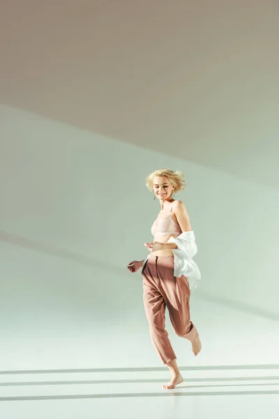 Smiling barefoot blonde woman in pink bra, shirt and pants running on grey — Stock Photo