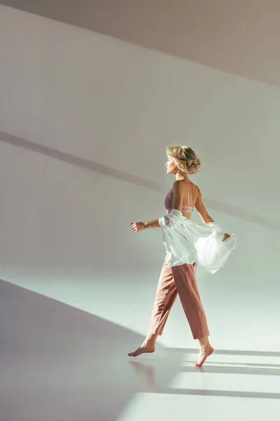 Vue latérale de fille blonde pieds nus en soutien-gorge rose, chemise et pantalon marchant sur gris — Photo de stock