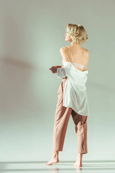 Back view of barefoot blonde girl in pink bra, shirt and pants standing and looking away on grey — Stock Photo
