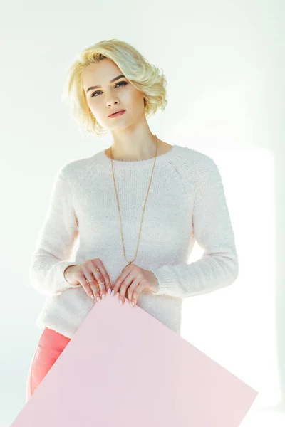 Belle fille blonde tenant une pancarte rose et regardant la caméra sur gris — Photo de stock