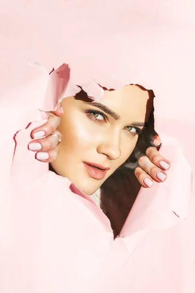 Красивая молодая женщина смотрит на камеру сквозь порванную розовую бумагу — стоковое фото