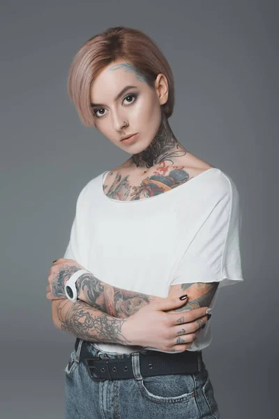 Portrait of beautiful girl with tattoos standing with crossed arms and looking at camera isolated on grey — Stock Photo