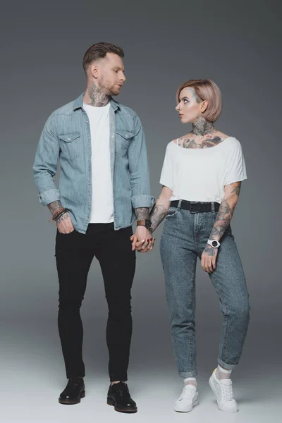 Full length view of beautiful stylish young tattooed couple holding hands and looking at each other on grey — Stock Photo