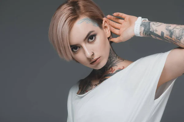 Retrato de chica tatuada atractiva en camiseta blanca mirando a la cámara aislada en gris - foto de stock