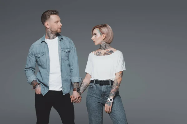Стильна молода пара з татуюваннями тримає руки і дивиться один на одного ізольовано на сірому — стокове фото