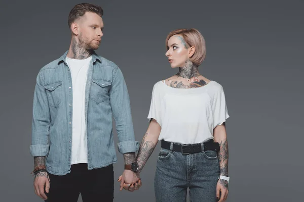 Schönes stylisches junges Paar mit Tätowierungen, die sich an den Händen halten und einander isoliert auf grau anschauen — Stockfoto