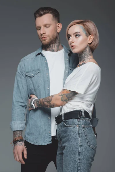 Hermosa pareja joven con estilo con tatuajes abrazos y mirando hacia otro lado aislado en gris - foto de stock