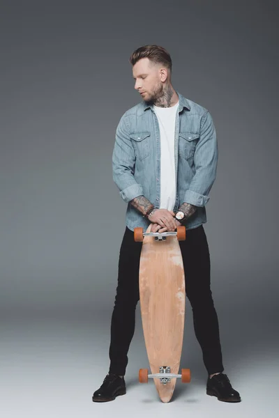 Vista completa di elegante giovane uomo tatuato in piedi con skateboard su grigio — Foto stock