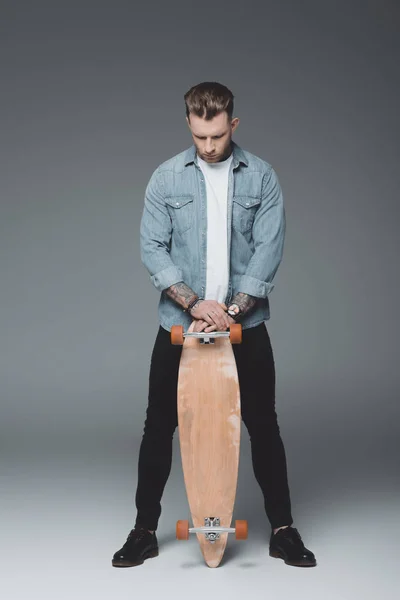 Piena vista di bel giovane tatuato in piedi con skateboard su grigio — Foto stock