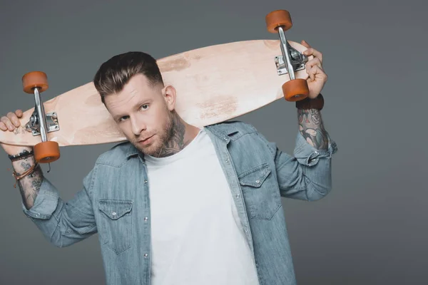 Стильний молодий татуйований чоловік тримає скейтборд на плечах і дивиться на камеру ізольовано на сірому — стокове фото