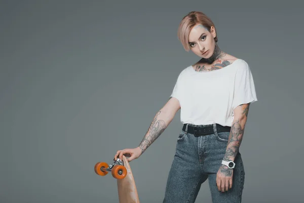 Красивая молодая татуированная женщина, стоящая со скейтбордом и смотрящая на камеру, изолированную на сером — стоковое фото