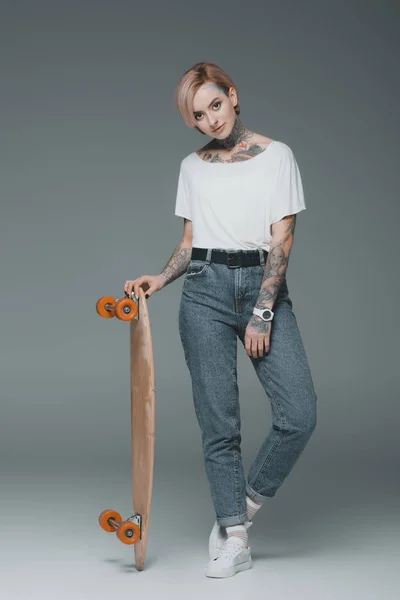 Hermosa chica tatuada de pie con monopatín y mirando a la cámara en gris - foto de stock