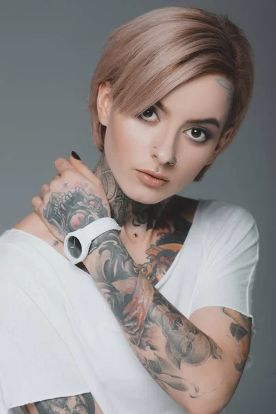 Retrato de hermosa chica tatuada en camiseta blanca mirando a la cámara aislada en gris - foto de stock