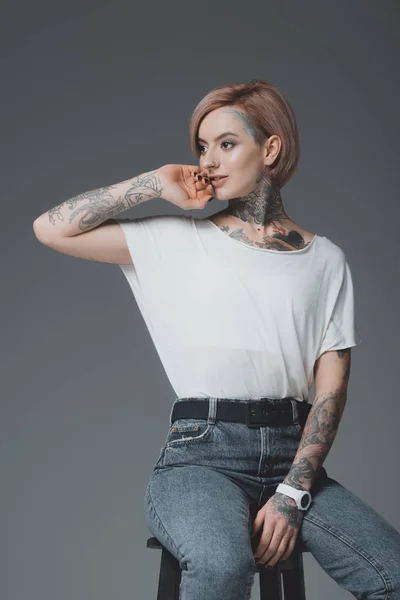 Hermosa joven sonriente con tatuajes sentado en el taburete y mirando hacia otro lado aislado en gris - foto de stock