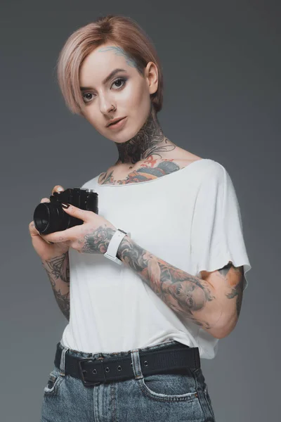 Hermosa joven tatuada mujer sosteniendo la cámara y mirando a la cámara aislada en gris - foto de stock