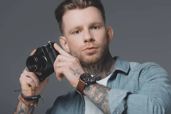 Красивий молодий чоловік з татуюваннями тримає камеру і дивиться на камеру ізольовано на сірому — стокове фото