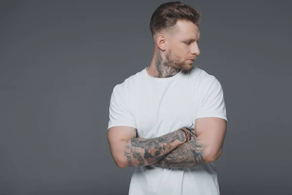 Elegante giovane uomo tatuato in t-shirt bianca in piedi con le braccia incrociate e distogliendo lo sguardo isolato sul grigio — Foto stock