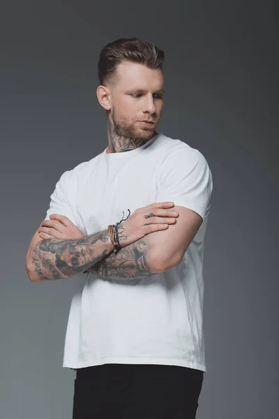 Bello elegante giovane uomo tatuato in bianco t-shirt in piedi con le braccia incrociate e guardando lontano isolato sul grigio — Foto stock