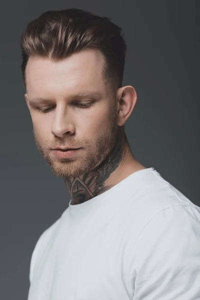 Retrato de guapo joven tatuado con estilo en camiseta blanca mirando hacia abajo aislado en gris - foto de stock