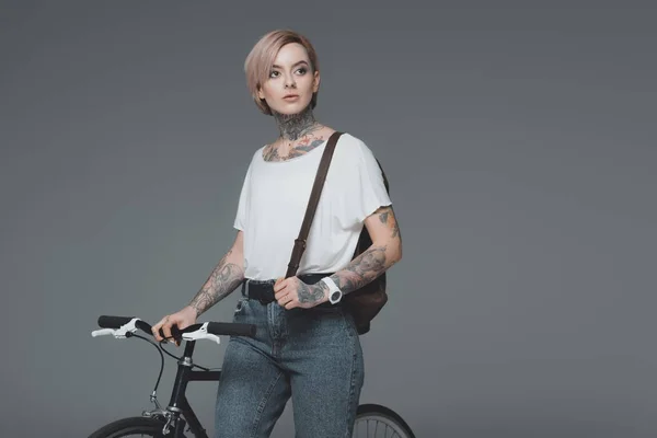 Красива татуйована дівчина з рюкзаком, що стоїть з велосипедом і дивиться ізольовано на сірому — стокове фото