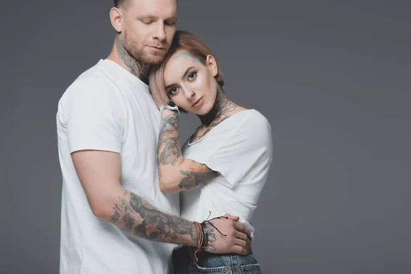 Elegante joven pareja tatuada en blanco camisetas abrazo aislado en gris - foto de stock