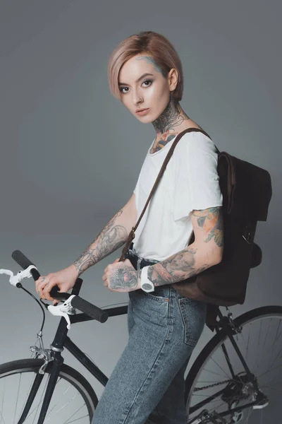 Beautiful tattooed girl with backpack standing with bicycle and looking at camera isolated on grey — Stock Photo