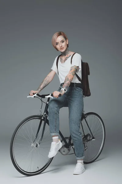Красива стильна дівчина з татуюваннями сидить на велосипеді і дивиться на камеру ізольовано на сірому — стокове фото