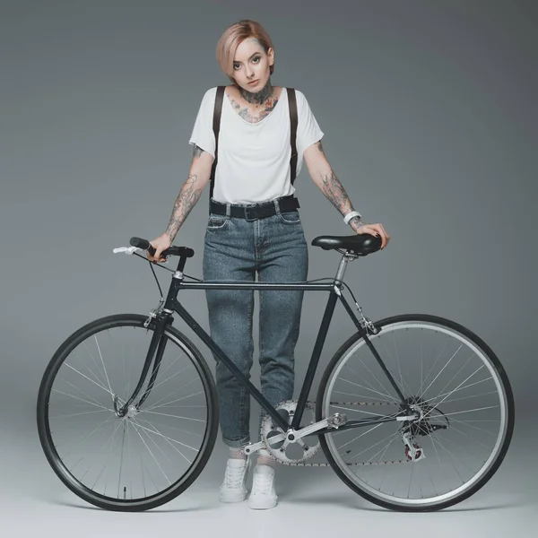 Hermosa chica elegante con tatuajes de pie con bicicleta y mirando a la cámara aislada en gris - foto de stock