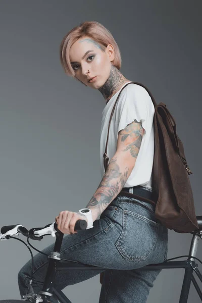 Hermosa chica tatuada sentada en bicicleta y mirando a la cámara aislada en gris - foto de stock