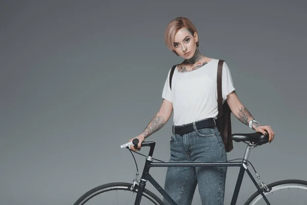 Красива татуйована дівчина стоїть з велосипедом і дивиться на камеру ізольовано на сірому — стокове фото