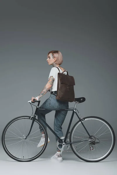 Elegante jovem tatuado mulher com mochila sentado na bicicleta e olhando para longe no cinza — Fotografia de Stock