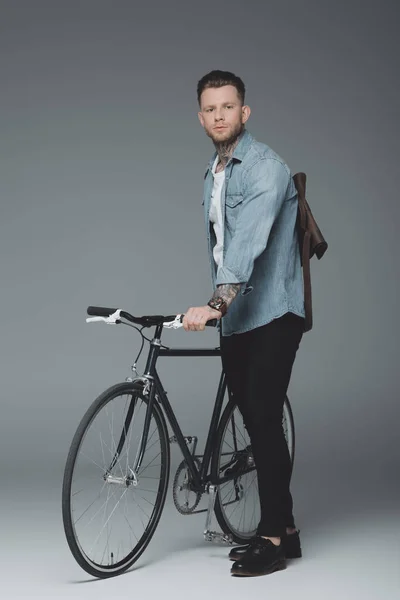 Vista completa de hombre joven con estilo con tatuajes de pie con bicicleta y mirando a la cámara en gris — Stock Photo