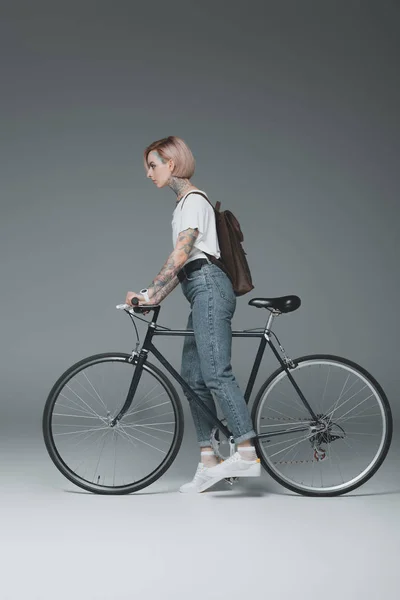 Side view of beautiful tattooed girl with backpack riding bicycle on grey — Stock Photo