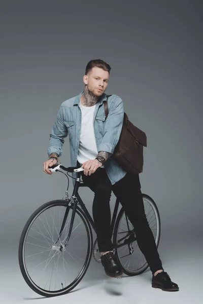 Élégant jeune homme tatoué assis sur le vélo et regardant la caméra sur gris — Photo de stock